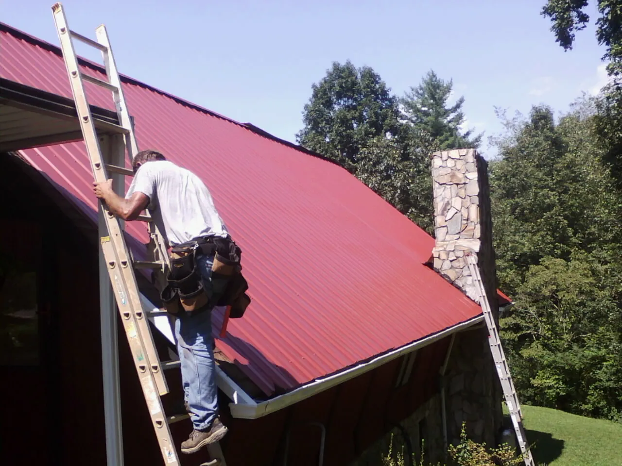 Roofer on a ladder beside a red metal roof and stone chimney — Soffit & Fascia Repair by Roofing Brothers in Orwigsburg